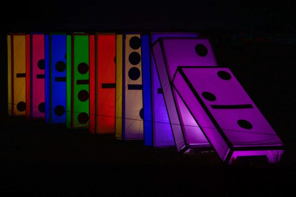 A row of luminous multicolored dominoes stand ready to tip over. The front two dominoes are leaning ready to cause a chain reaction.