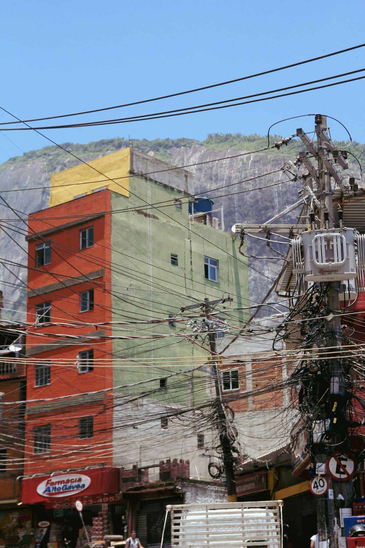 Two wooden power poles bedecked with hundreds of different wires stand in front of old brick buildings up to six stories high. In the background is a tall grey sheer cliff and blue sky.