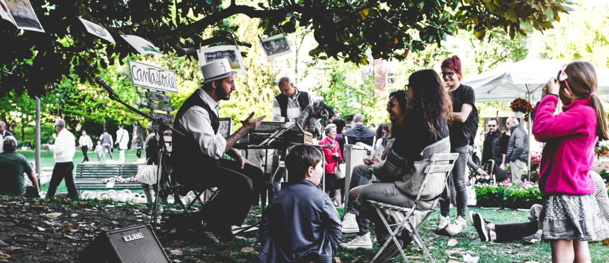 A group of children sit under a tree listening to a person in a waistcoat and hat tell a story.