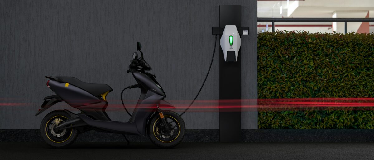 An electric scooter is parked in front of a concrete wall and plugged into a charger. A red line in the front of the image is a long exposure of a vehicle with its lights on passing by.
