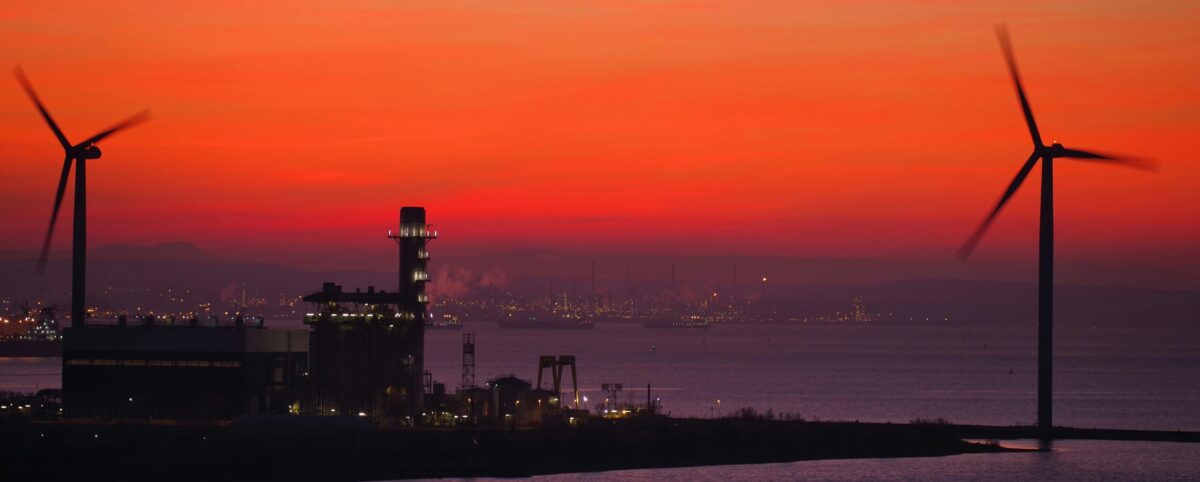 A vivid sunrise with bright orange and red sky is the background for two wind turbines powering a port with many large container ships in silhouette