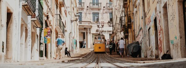 A yellow tram in the distance at the end of electric tram lines and overhead wires, at the sides of the pictures are glowing white buildings from centuries past and some pedestrians