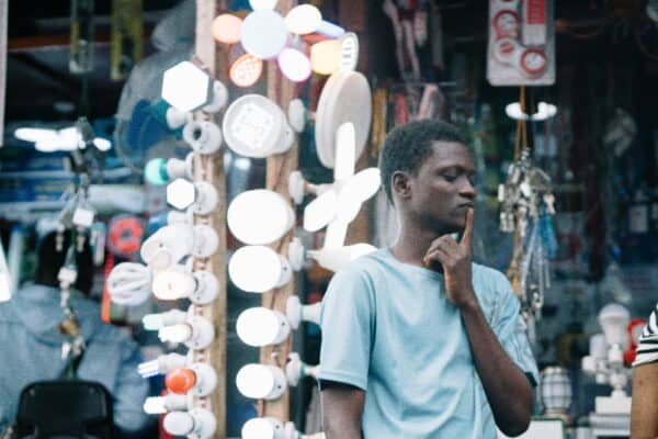 Young Man Thinking in Market A young black man stands with his eyes closed and one fingerto his lips, thinking in front of a stand of various electric lightbulbs